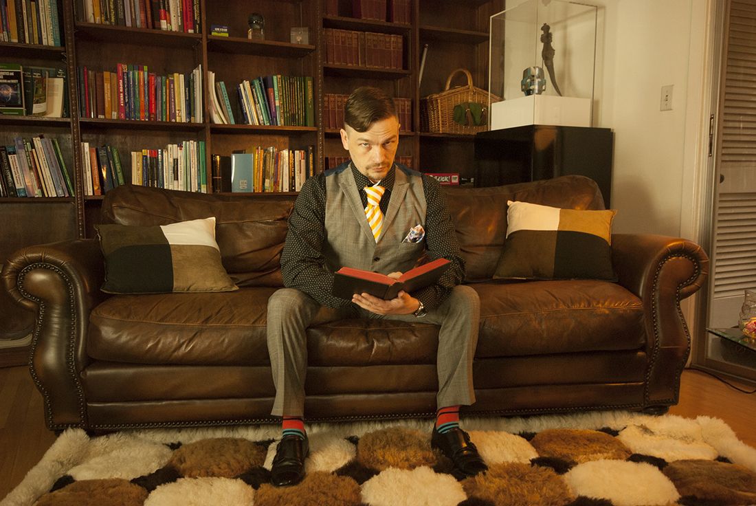 Josh Pritchard seated on a leather couch in front of a wall of bookshelves, reading.